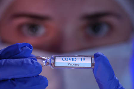 A doctor in a medical mask and blue sterile gloves holds a glass ampoule with a coronavirus vaccine...