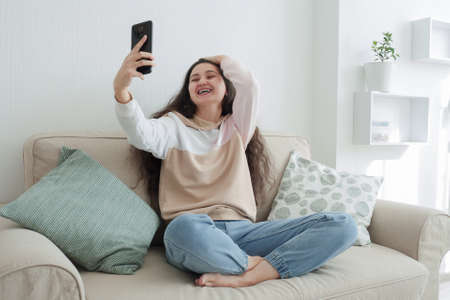 A young Asian girl with long curly hair in blue jeans and a beige hoodie communicates with friends via video chat on a smartphone and laughs cheerfully. Online communication conceptの写真素材