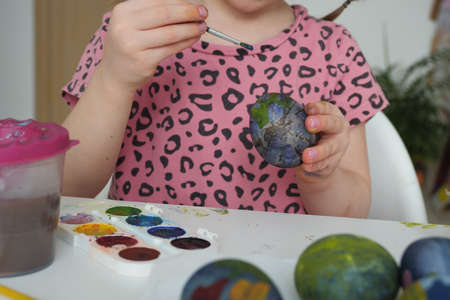 A child girl sits at home at the table and paints Easter eggs with watercolors for Easter and for playing. Homemade Easter traditions of painting eggsの写真素材