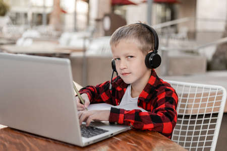A bright boy, a schoolboy, a student in a checkered red shirt, sits in the school yard at a table with headphones and a laptop. Watching video and yelling at the screen, or talking via video call. distance learning. homeschooling. An elementary school student won the competition.の写真素材