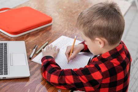 A bright boy, a schoolboy, a student in a checkered red shirt, sits in the school yard at a table with headphones and a laptop. Watching video and yelling at the screen, or talking via video call. distance learning. homeschooling. An elementary school student won the competition.の写真素材