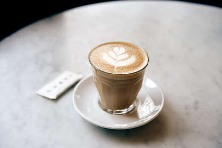 A cup of coffee with latte art on the white round marble table. A sugar sachet is near the cup.の写真素材