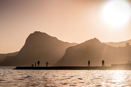 Silhouettes of people and rocks against the setting golden sun on the black sea. Shaft of lights make Silhouette. Shadow at sunset. Black sea beach in Sudak, Crimea.の写真素材