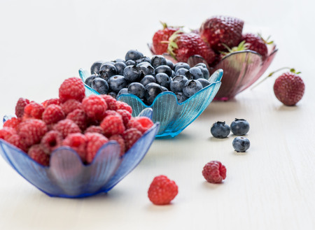 Servings of strawberries, blueberries and raspberries in colored bowls on a white tableの写真素材