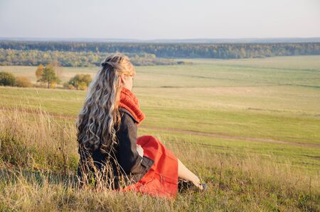 A lonely girl with long curly hair sitting on a hill in front of a large meadowの写真素材