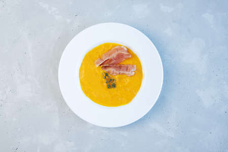 Pumpkin puree soup with bacon and a sprig of thyme in a white ceramic plate on a gray backgroundの写真素材
