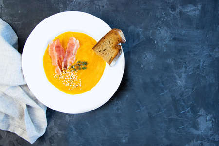 Pumpkin soup with bacon decorated with hemp seeds and a sprig of rosemary, toasted piece of bread on a plate. Next to a linen table napkin. Dark background. Place for text.の写真素材