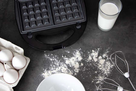 making wafers at home - a glass of milk, a waffle maker, eggs, flour and a whisk for mixing. Cooking backgroundの写真素材