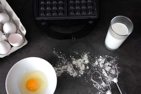 making waffles at home - a glass of milk, a waffle maker, a broken egg in a bowl, flour and a whisk for mixing. cooking Viennese waffles. Place for textの写真素材