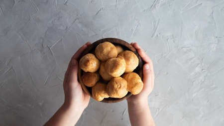 Hands holding a small wooden bowl with homemade eclairs over a gray table. Plate with small round eclairs in the hands. Place for text.の写真素材