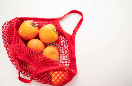 A red cotton string bag with persimmons and tangerines lies on a white background. The concept of...