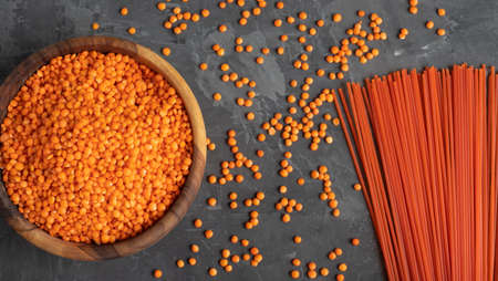 Red lentils in a wooden bowl, next to scattered grains and pasta from lentils on a dark background....
