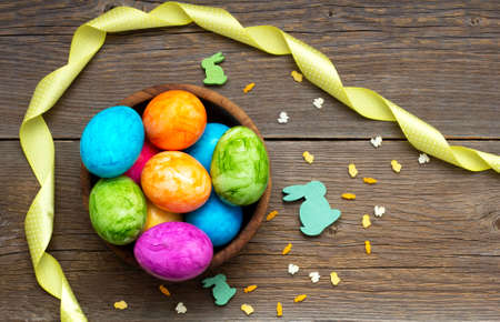 Easter flat lay. Colorful eggs lie in a wooden bowl on an old rustic table. Nearby there is a twisted yellow ribbon and wooden decorative hares.の写真素材