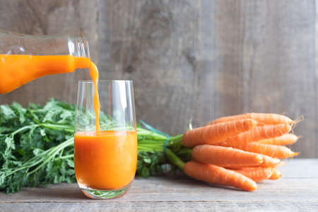Carrot juice is poured from a glass bottle into a glass, a bunch of fresh carrots with green tops lies at the back. Dietary nutrition. Healthy lifestyle. Wood background.の写真素材