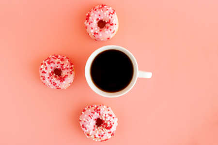 Top view of a cup of black coffee and around are three donuts with pink icing on a pink background. Place for text. Pause for coffee. Snack.の写真素材