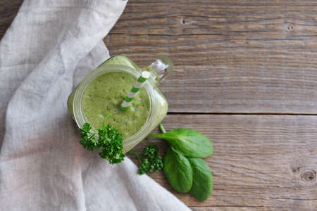 Top view on a glass of green smoothie and spinach leaves on a wooden background, linen table napkin...
