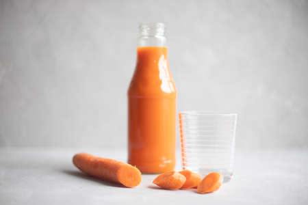 Bottle with fresh carrot juice and an empty glass nearby. In the foreground is a carrot. Grey background. Place for text.の写真素材