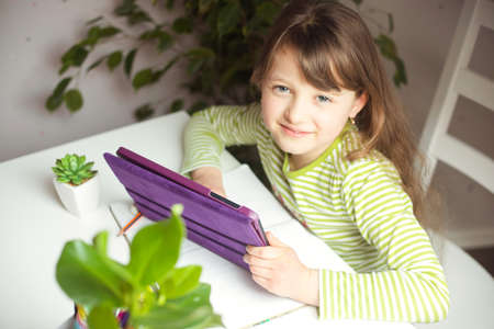 A cute girl, a European-style girl, sits at a table in front of a tablet and is engaged, doing homework. Home schooling.の写真素材