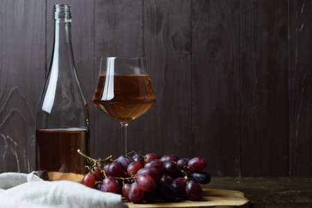 An open bottle of wine next to a glass of pink wine, a bunch of red grapes and a linen table napkin on a wooden board on a wooden background. Place for text.の写真素材
