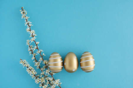 Three golden easter eggs painted with white paint lie on a blue background. Nearby lies a branch with white flowers. Easter concept. Place for textの写真素材