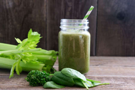A glass with a green smoothie and a bamboo cocktail straw is standing on a wooden board. Nearby are bunches of celery and spinach and parsley leaves. Useful snack. Vegan, vegetarian drink. Copy space.の写真素材