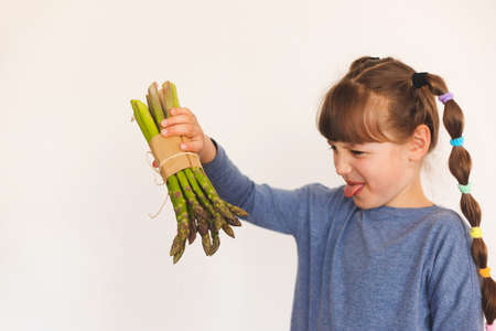 A girl with two ponytails holds a bunch of asparagus on her outstretched hand and makes a face. Children do not like vegetables. White background. Place for text.の写真素材