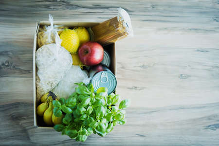 Cardboard box with various essentials, cereals, canned food, pasta, bananas, apples on a wooden table. Place for text.の写真素材