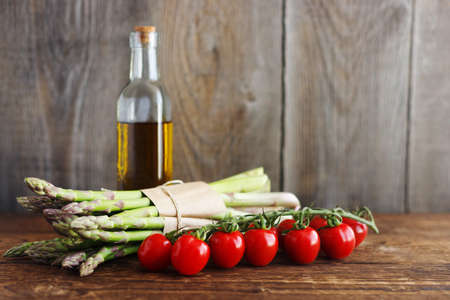 A bunch of fresh green asparagus lies next to a branch of cherry tomatoes, in the background is a glass bottle with olive oil. Wooden background, place for text.の写真素材