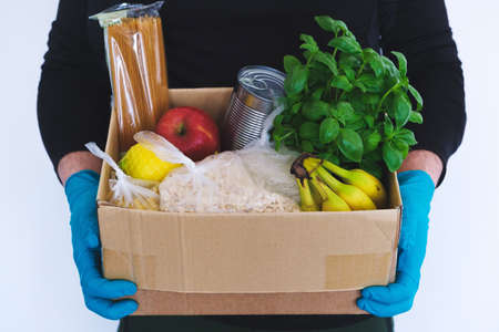 Male hands in medical gloves hold a cardboard box with products of oatmeal, rice, canned food, pasta, apples, lemons. Food donation, contactless deliveryの写真素材