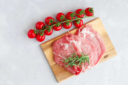 Raw pork steaks lie on a wooden cutting board with a sprig of rosemary and next to them are red cherry tomatoes on a branch. Grey background. Copy space.の写真素材