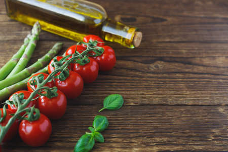 Cherry tomatoes on a branch, green asparagus and a bottle of olive oil lie on a wooden old table. Preparation of ingredients for cooking. Copy space.の写真素材