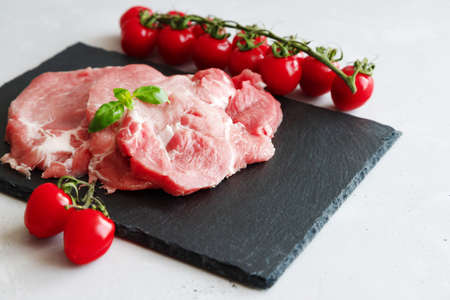 Raw fresh pork steaks with a sprig of basil and cherry tomatoes on a branch lie on a black stone cutting board. Preparing meat for cookingの写真素材