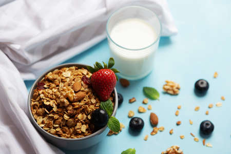 A bowl, a cup with freshly prepared granola is on a blue table next to a glass of milk. Homemade granola with fresh strawberries and blueberries. Proper nutritionの写真素材