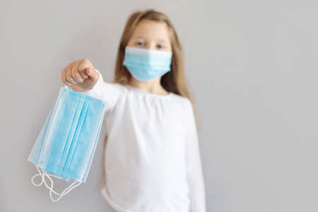 Cute serious girl with long blond hair in a mask, out of focus, holds medical masks on an outstretched arm. Virus protection. Place for text.の写真素材