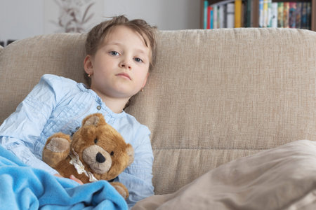 A sad or sick girl with brown hair of European appearance sits on a beige sofa at home. Childhood depression, lack of understanding of adults, conflicts with parents.の写真素材