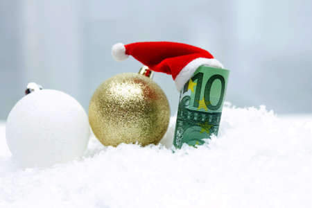 A roll of banknotes of 100 euros in a New Years cap of Santa Claus lie in white snow next to Christmas balls of different colors.の写真素材