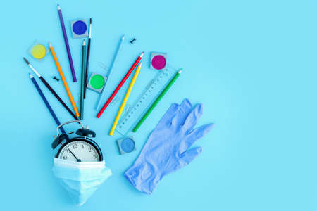 Stationery, crayons, brushes, medical mask and a glove around a black alarm clock. Back to school,...