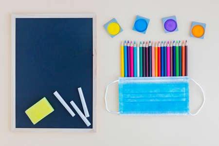 Colored pencils and brushes under a protective medical mask and next to a graphite board with chalk on a pink background with space for text. Post-coronavirus training. Back to schoolの写真素材
