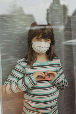 A cute girl schoolgirl stands outside the window and makes a heart gesture with her hands. Coronovirus. Vertical orientation.の写真素材