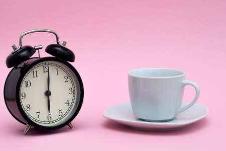 A black alarm clock in retro style and a white coffee cup on a small saucer stand on a pink background. Good morning concept. Morning coffeeの写真素材