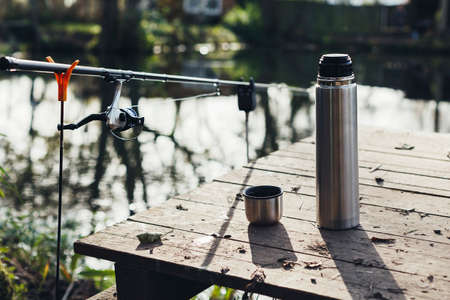 A tumbler with hot tea, coffee is on a wooden deck on the shore of the lake and next to a fishing rod. Travel alone, outdoor recreation, halt, rest, fishing.の写真素材