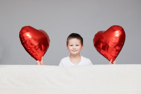 Valentines Day, a holiday for all lovers. A cute Caucasian boy with red balloons peeks out from behind the sofa. Copy space. Congratulations to loved onesの写真素材