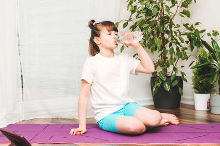 A cute Caucasian girl is engaged in drinking water from a bottle during breaks from sports. Fitness at home, the importance of water in sports.の写真素材