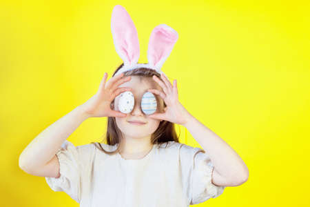 Easter concept. Portrait of a cute girl with bunny ears dabbles with painted eggs, covers her face...