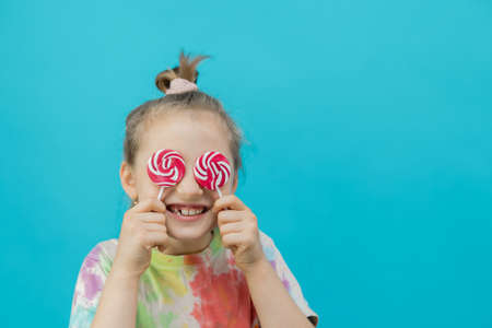 The concept of a holiday, fun entertainment and childrens joy. A cute dark-haired girl in a multi-colored T-shirt holds round lollipops and closes her eyes with them. Close-upの写真素材