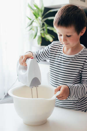 A charming cheerful boy kneads dough with a mixer in the kitchen. Children cat dough at home. Homemade baking. Authentic moments of lifeの写真素材