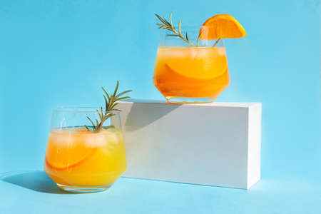 Lemonade, orange cocktail, refreshing summer drink with ice and orange slices in a glass with a sprig of rosemary on a white podium. Trending drinks.の写真素材