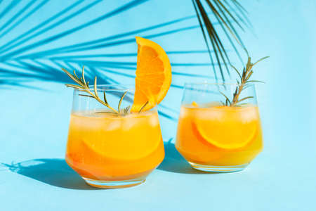 Close-up lemonade, orange cocktail, refreshing summer drink with ice in a glass with a sprig of rosemary on a blue background under a palm leaf on a bright sunny day.の写真素材