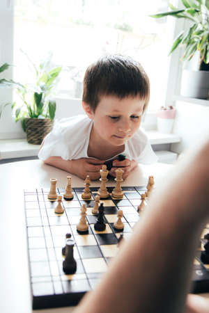 A cute Caucasian boy is thinking about the next move in a board game. Chess for the development of logic and thinking in children. Vertical photoの写真素材