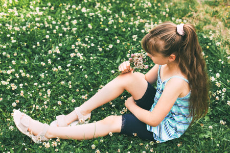 A cute Caucasian girl is sitting in a clover meadow and collecting a bouquet. Rest from the city....